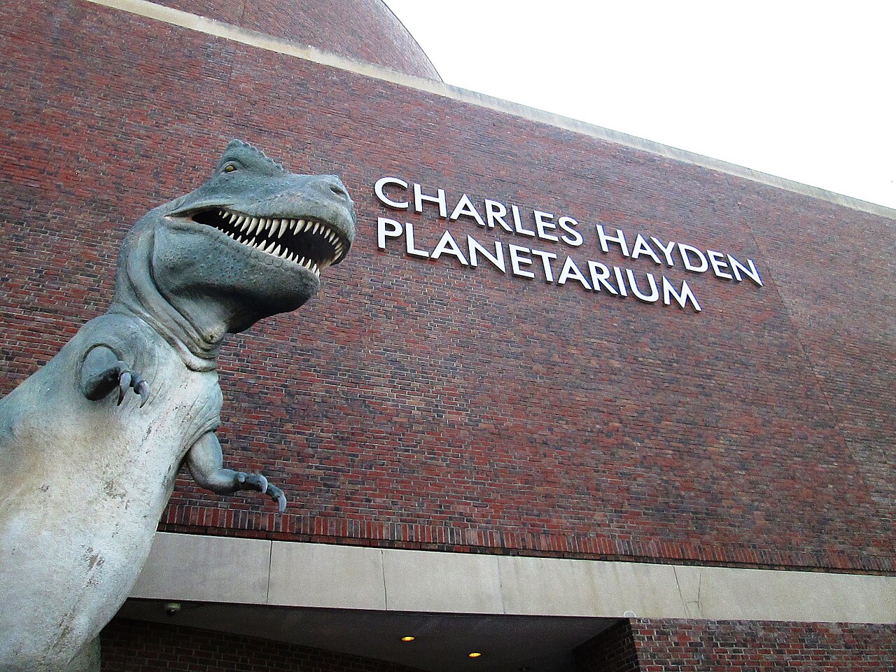 A dinosaur model beside the exterior Charles Hayden Planetarium entrance sign in Boston, photographed in 2017